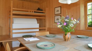 A wooden interior with a dining table, chairs, a vase with flowers, and shelves with bedding.