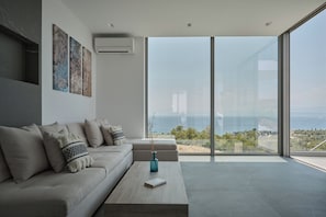 A modern living room with a sofa, a coffee table, and a view through large glass doors. - Villa Anthos | Sea-view Villa with Pool & Jacuzzi (Βερβεροντα,)