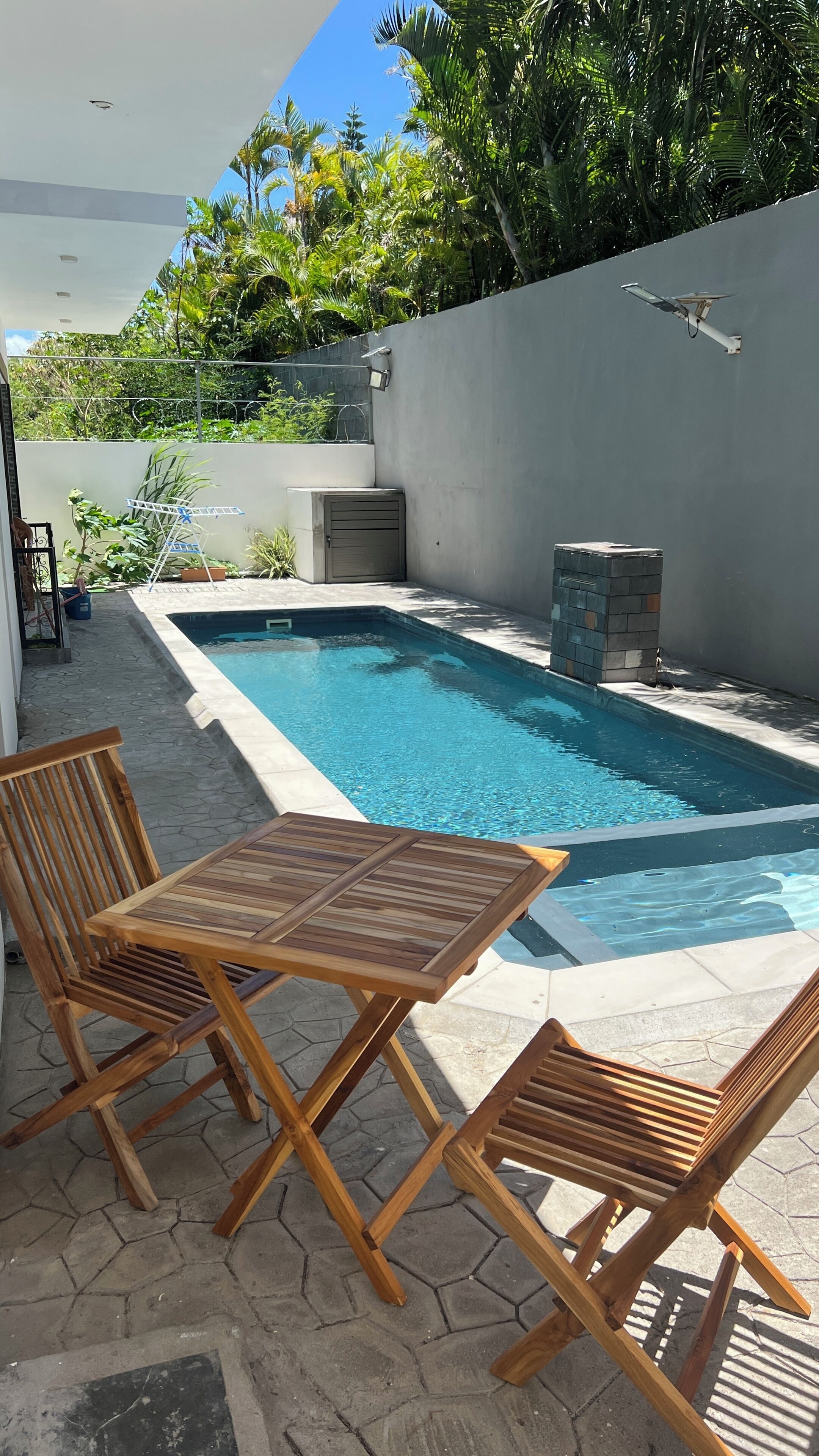 A swimming pool with a tiled deck, surrounded by a low wall and lush greenery.