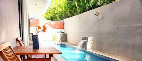 Una piscina con una pequeña cascada, rodeada por un patio de piedra y muebles de madera.