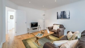 Uma sala de estar moderna com uma mesa de centro de madeira, um sofá de couro marrom, um rack branco para TV e uma pintura com vista para a cidade na parede.
