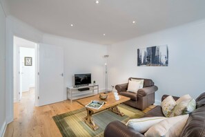 A modern living room with a wooden coffee table, a brown leather sofa, a white TV stand, and a cityscape painting on the wall. - Urban Retreat in Vauxhall - 2BR Central Stay (London)