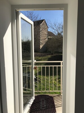 Blick durch eine Glastür auf einen Balkon mit Metallgeländer, ein Steinhaus im Hintergrund und ein klarer Himmel.
