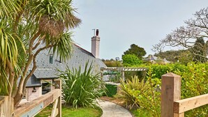 A pathway leading to a house with a chimney, surrounded by lush greenery and a wooden fence. - Rosedale Cottage (Brixham)