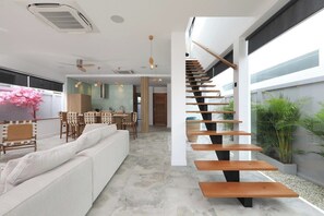 A modern interior with a staircase, a sofa, dining area, and a view of greenery. - Modern 4BR Villa with Pool | Rawai | Capital Pro (Rawai)