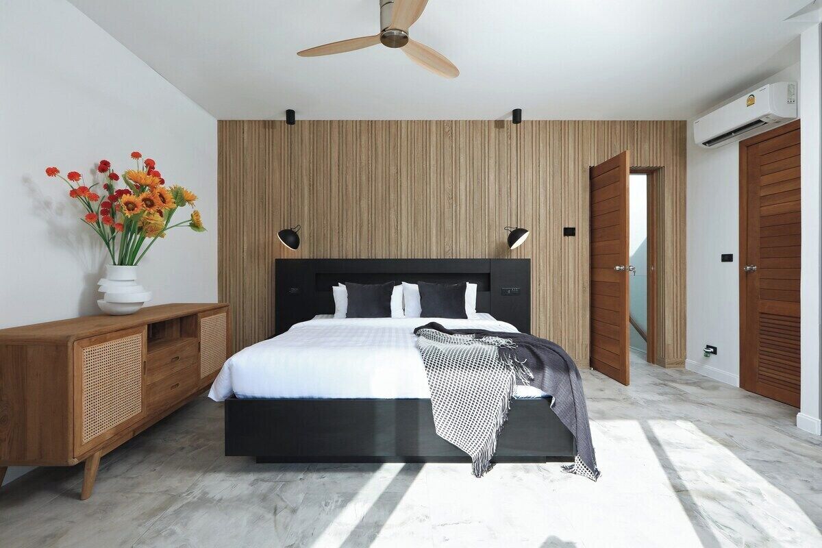 A modern bedroom with a bed, wooden panel wall, ceiling fan, bedside lamps, and a wooden wardrobe.