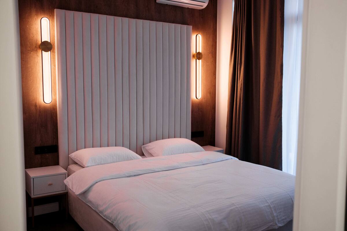 A modern hotel room with a large bed, two bedside tables, a headboard with vertical lines, and wall-mounted lighting.