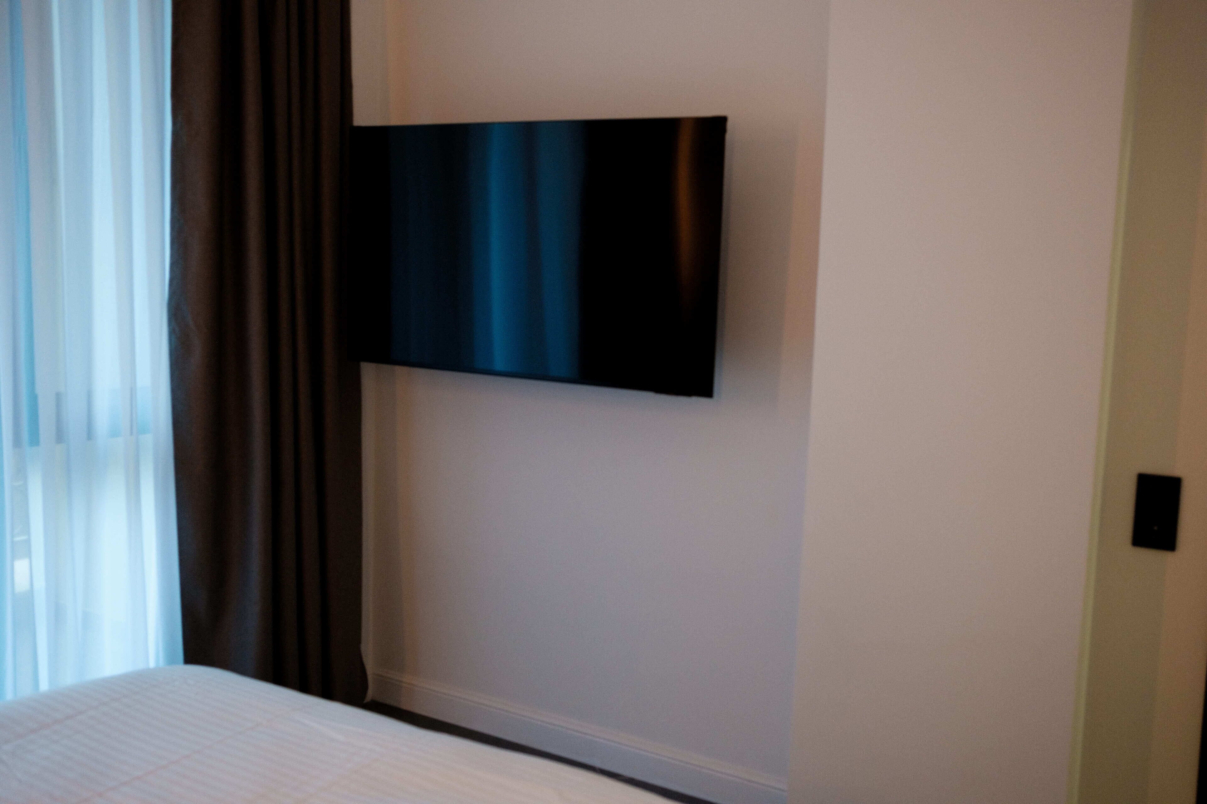 A hotel room with a bed, a flat-screen TV mounted on the wall, and a window with curtains.