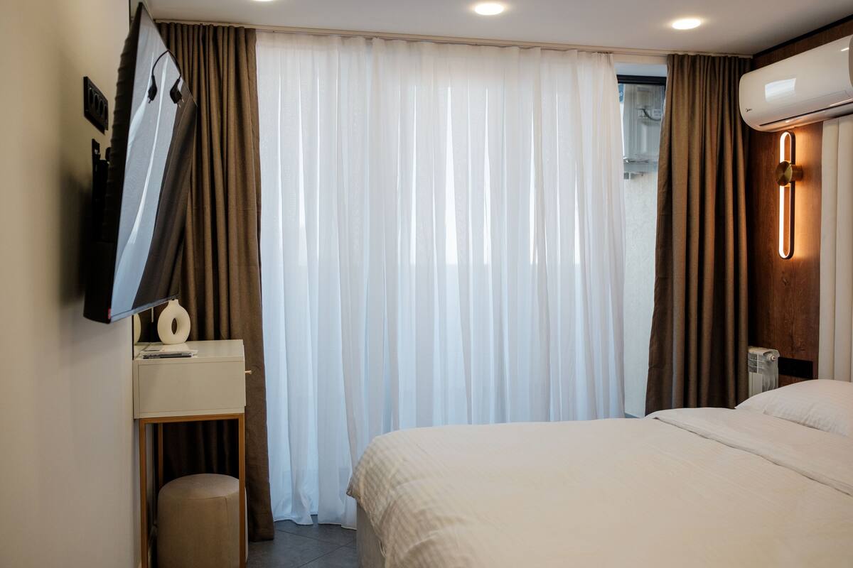 A hotel room with a bed, a desk with a lamp, a mirror, and a wall-mounted air conditioner.