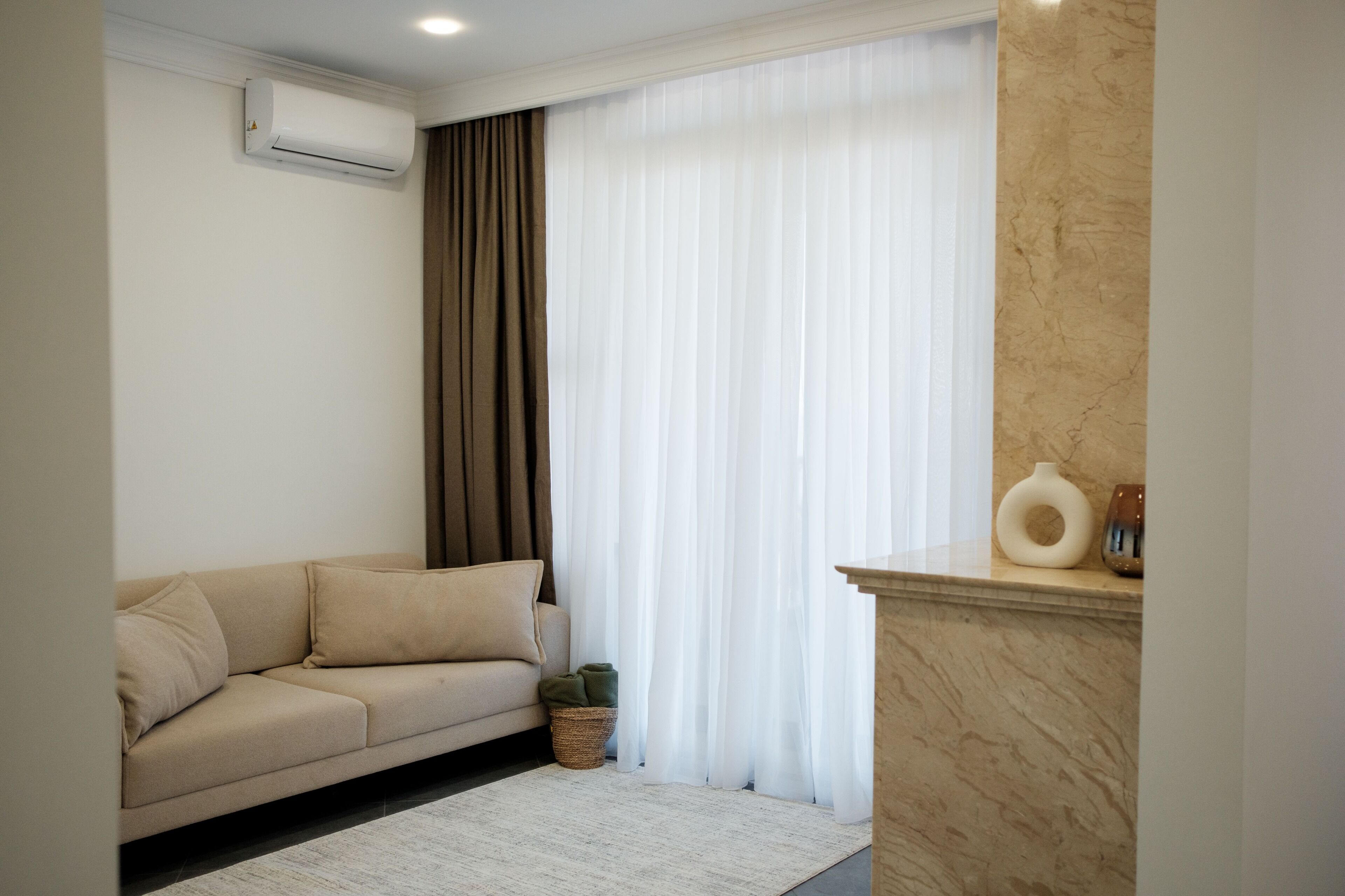 A living room with a beige sofa, a white air conditioner, a window with sheer white curtains, a marble fireplace, and a small basket with a plant.