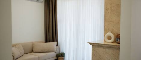 A living room with a beige sofa, a white air conditioner, a window with sheer white curtains, a marble fireplace, and a small basket with a plant.