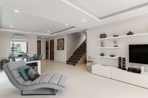 A modern living room with a grey sofa, a staircase, a flat-screen TV, and a minimalist decor. - Laguna Park | Bangtao Beach | 5-bedroom Rooftop Pool Villa (Tambon Choeng Thale)