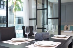 A dining table set with plates, cutlery, and wine glasses, with a view of a modern building and a palm tree outside. - 2BR Apartment · Palmyrah Surin · Surin | CapitalPro (Tambon Choeng Thale)