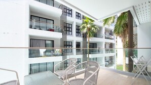 Ein Balkon mit Korbmöbeln, einer Glastrennwand und Blick auf ein Gebäude mit mehreren Balkonen und einer Palme.