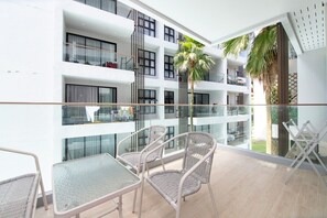 Een balkon met rieten meubels, een glazen hekwerk en uitzicht op een gebouw met meerdere balkons en een palmboom.