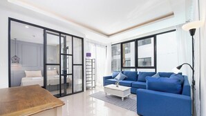 Ein modernes Wohnzimmer mit einem blauen Sofa, einem Holztisch und einer Glastür zum Schlafzimmer.