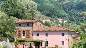 Una casa in pietra a due piani con facciata rosa e tetto in tegole rosse, circondata da vegetazione rigogliosa e colline.