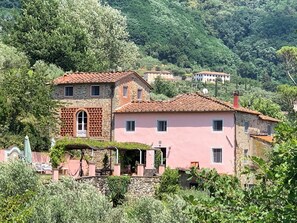 A two-story stone house with a pink facade and a red-tiled roof, surrounded by lush greenery and hills. - Apartment in farmhouse, nestled in olive grove, 15 minutes from Lucca (Capannori)