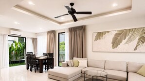 A modern living room with a sectional sofa, a dining table with chairs, a ceiling fan, and a large window with curtains. - 3BR Townhouse with Jacuzzi & Lake View |CapitalPro (Tambon Choeng Thale)