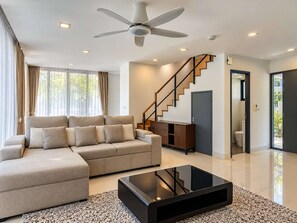 Un salon moderne avec un canapé d’angle, une table basse en verre, un ventilateur de plafond et un escalier menant à un autre niveau.