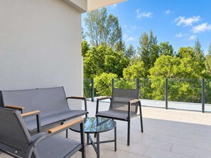 Un balcon avec un canapé et deux fauteuils, une table en verre et une vue sur les arbres et le ciel.