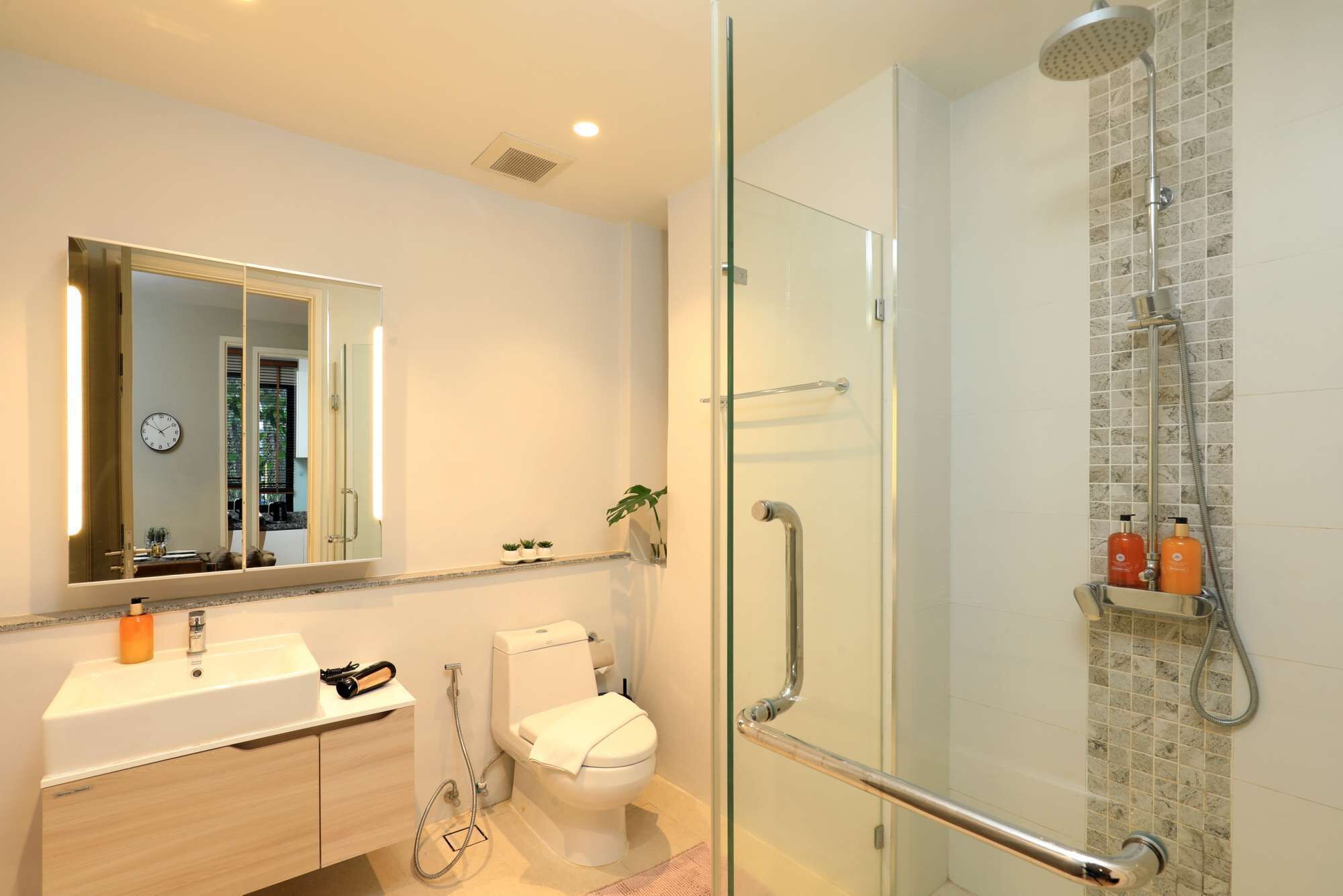 A modern bathroom with a glass shower enclosure, a white toilet, a sink with a mirror, and a clock on the wall.