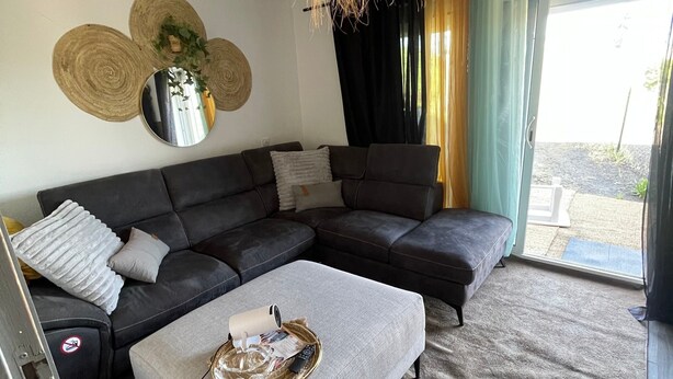 En stue med en hjørnesofa, et rundt spejl, en vævet loftlampe og udsigt gennem en glasdør.