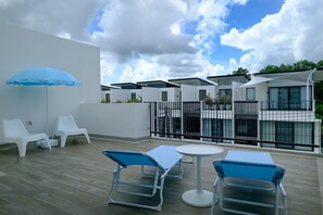Eine Dachterrasse mit weißen Liegestühlen, einem blauen Sonnenschirm und einem weißen Tisch.