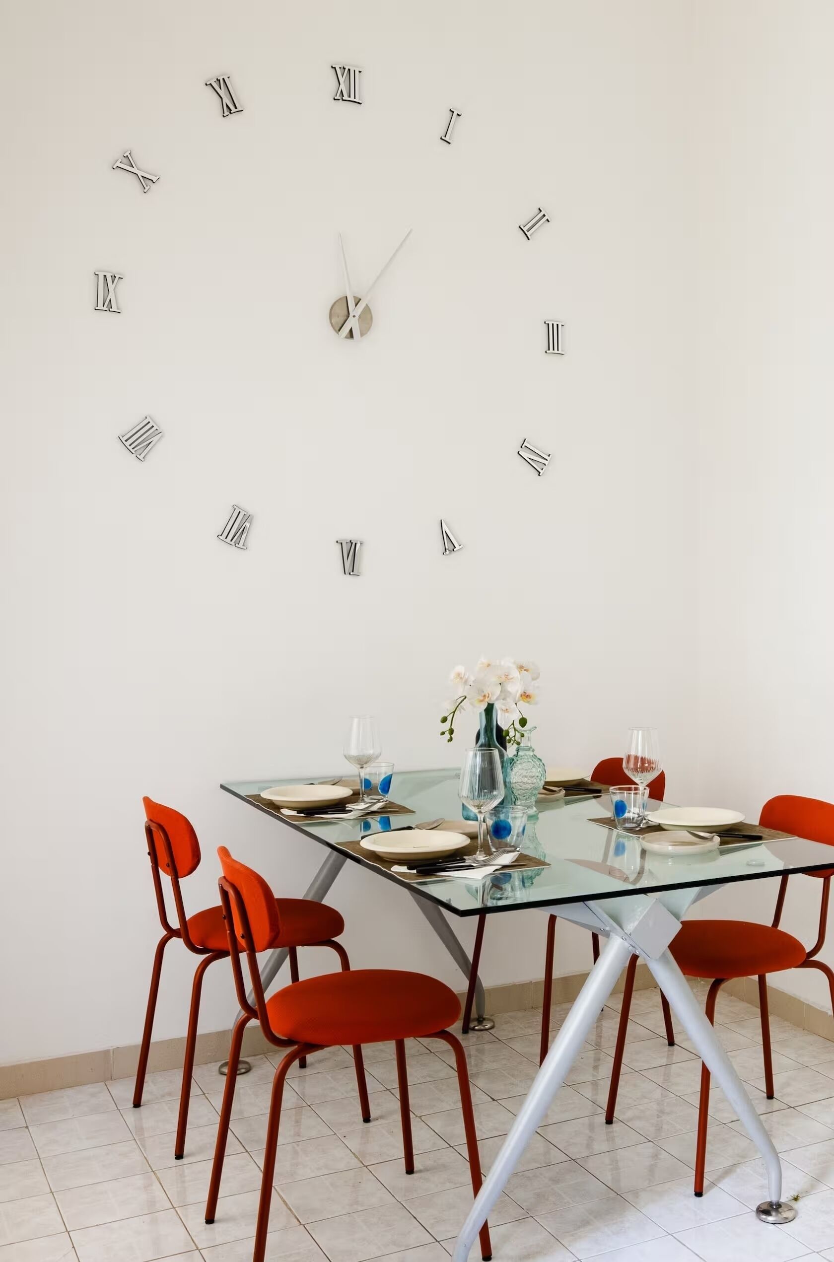 Zona pranzo con tavolo in vetro, sedie rosse, orologio a parete e un vaso con fiori.