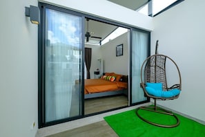 A room with a bed, a wicker chair, a green rug, and a sliding glass door leading to another room. - 3BR Townhouse Near Bangtao Beach | Capital Pro (Tambon Choeng Thale)