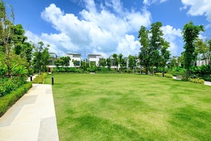 A well-maintained grassy area with a paved walkway, surrounded by trees and modern buildings. - 3BR Townhouse · Laguna Park · Bangtao | CapitalPro (Tambon Choeng Thale)