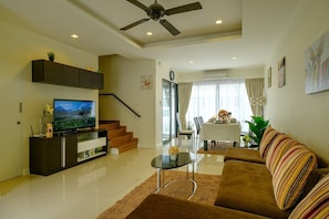 A modern living room with a flat-screen TV, a sofa, a coffee table, a staircase, and a dining area with a table and chairs. - 3BR Townhouse · Laguna Park · Bangtao | CapitalPro (Tambon Choeng Thale)