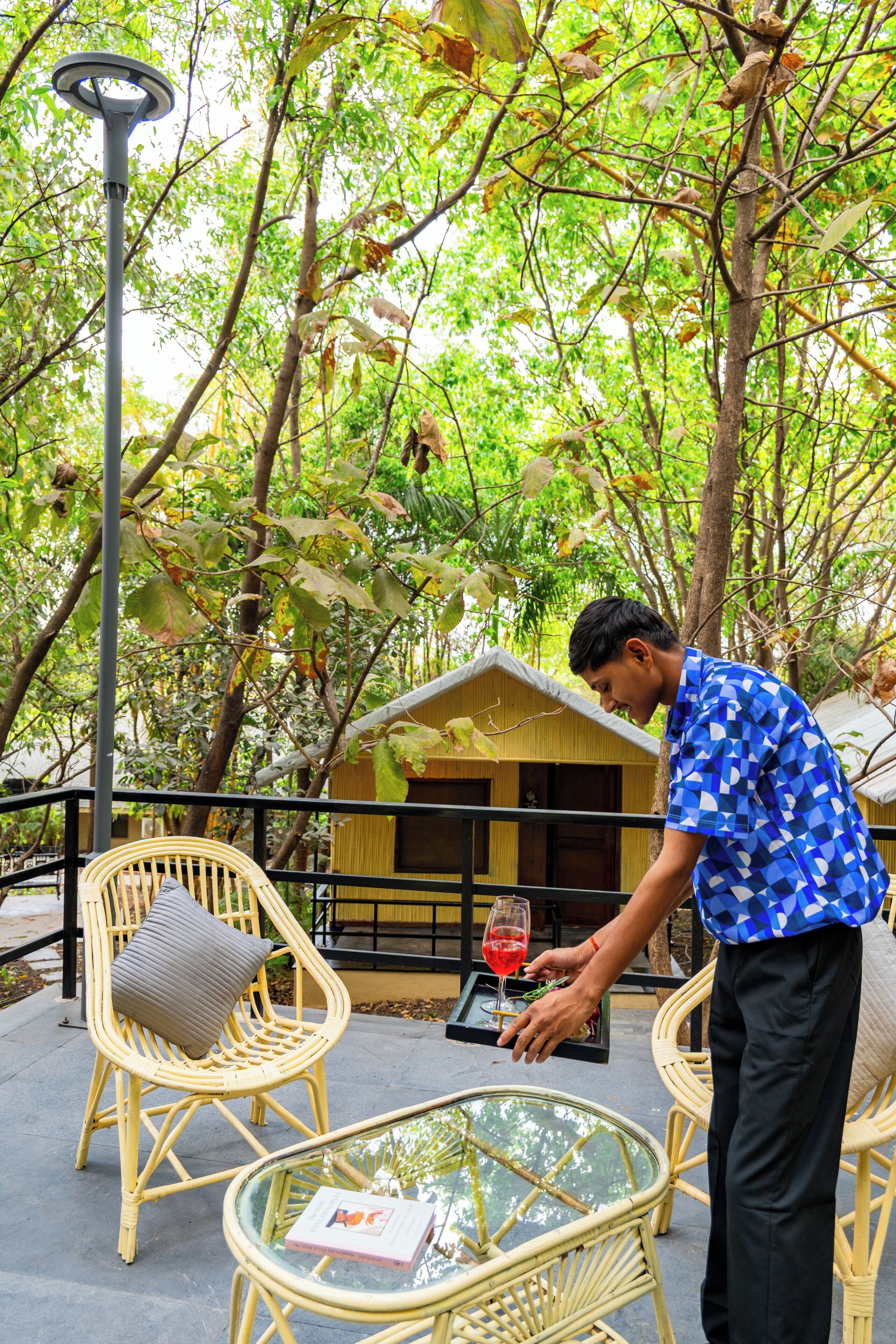 Un homme sert une boisson à une table en verre, entourée de chaises en osier, avec un petit bâtiment en arrière-plan.