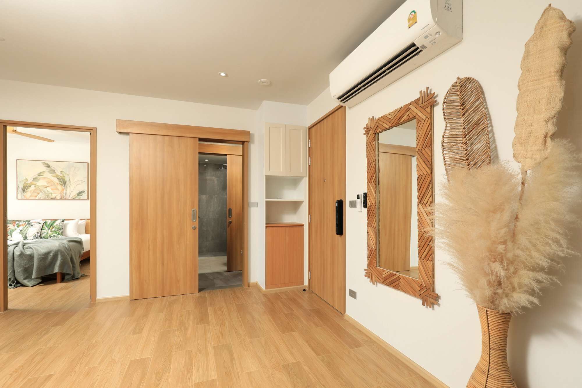 A hotel room with wooden flooring, a bed with a green blanket, a wardrobe, a bathroom with a shower, and a wall-mounted air conditioner.