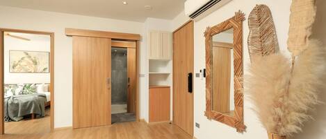 A hotel room with wooden flooring, a bed with a green blanket, a wardrobe, a bathroom with a shower, and a wall-mounted air conditioner.
