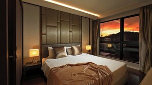 Habitación de hotel con una cama grande, dos lámparas de noche, vistas a un atardecer y una ventana con cortinas.