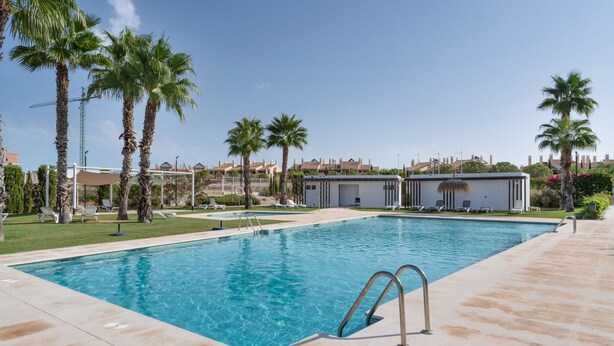 Une piscine rectangulaire à l’eau d’un bleu limpide, entourée d’une terrasse carrelée. On aperçoit des palmiers et un bâtiment à toit plat à l’arrière-plan.