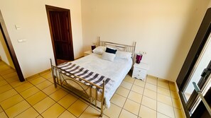 Un llit individual amb estructura metàl·lica, una taula de nit amb una làmpada, i una porta al fons.