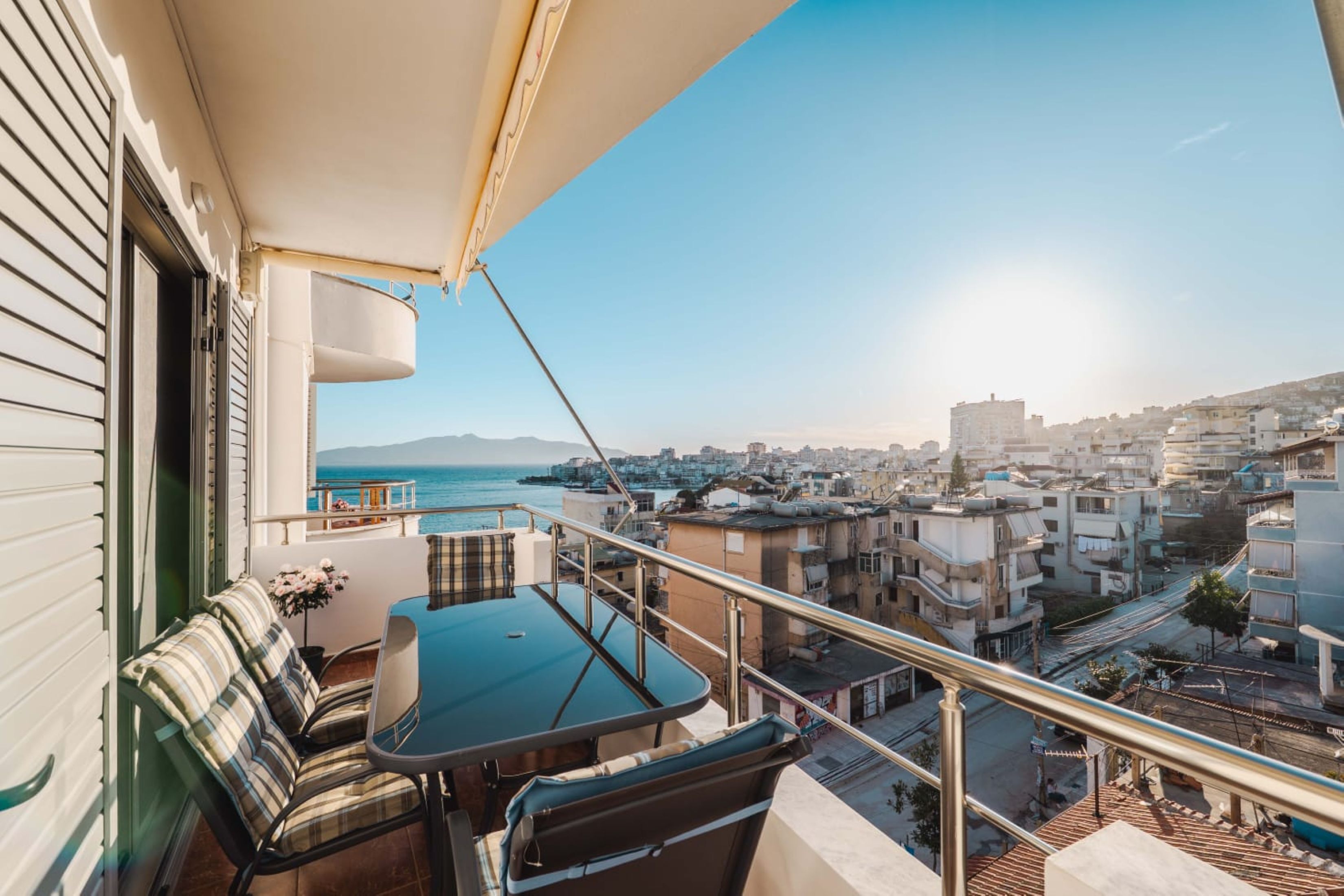 A balcony with a table and chairs, overlooking a cityscape and the sea.