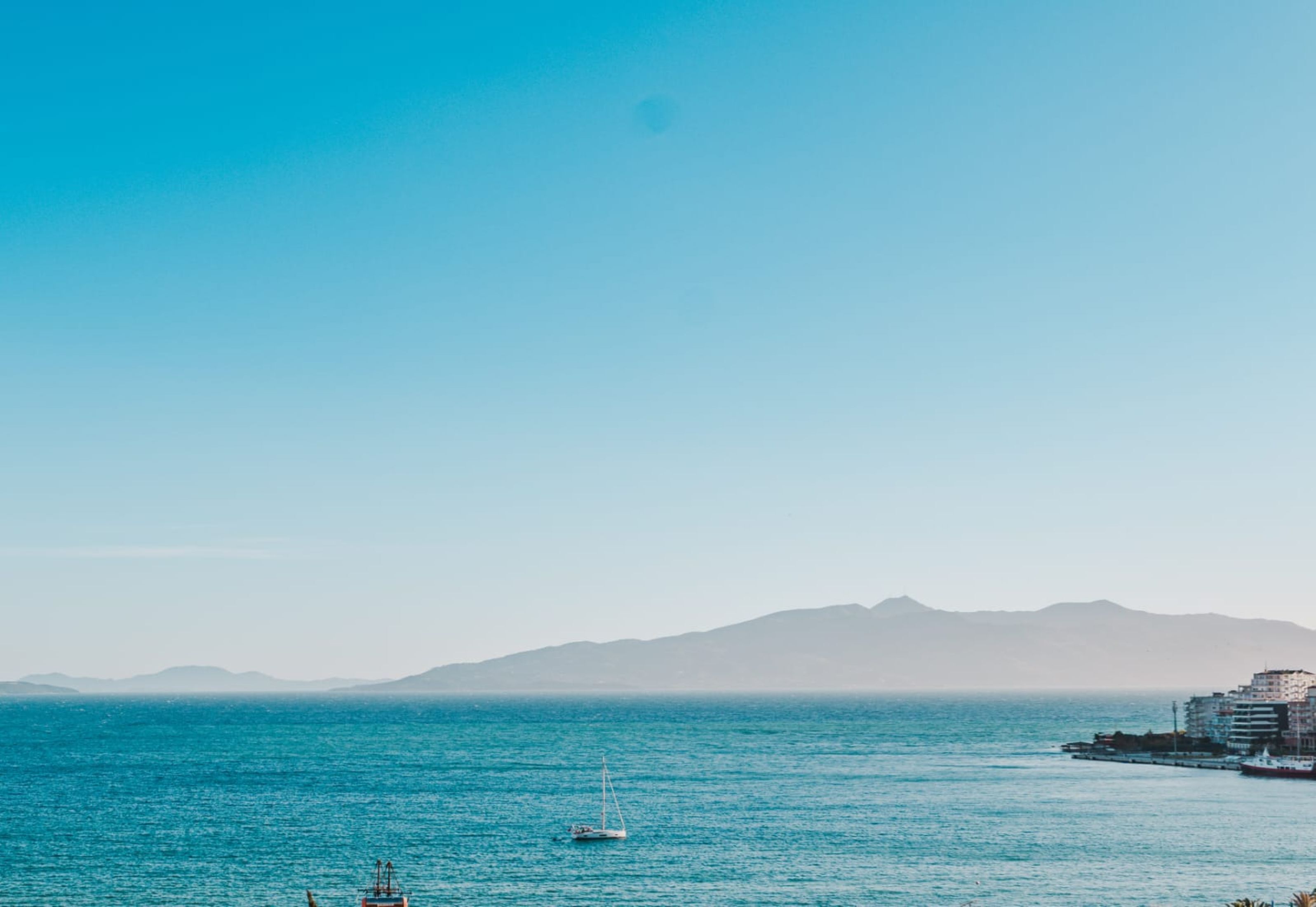 Vista costera con un velero, mar tranquilo y montañas lejanas.