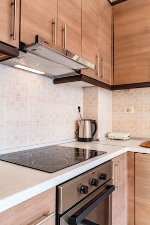 A kitchen with wooden cabinets, a stainless steel range hood, a built-in induction cooktop, an oven, and a kettle on the countertop. - Albania Dream Saranda Apartments (Saranda)