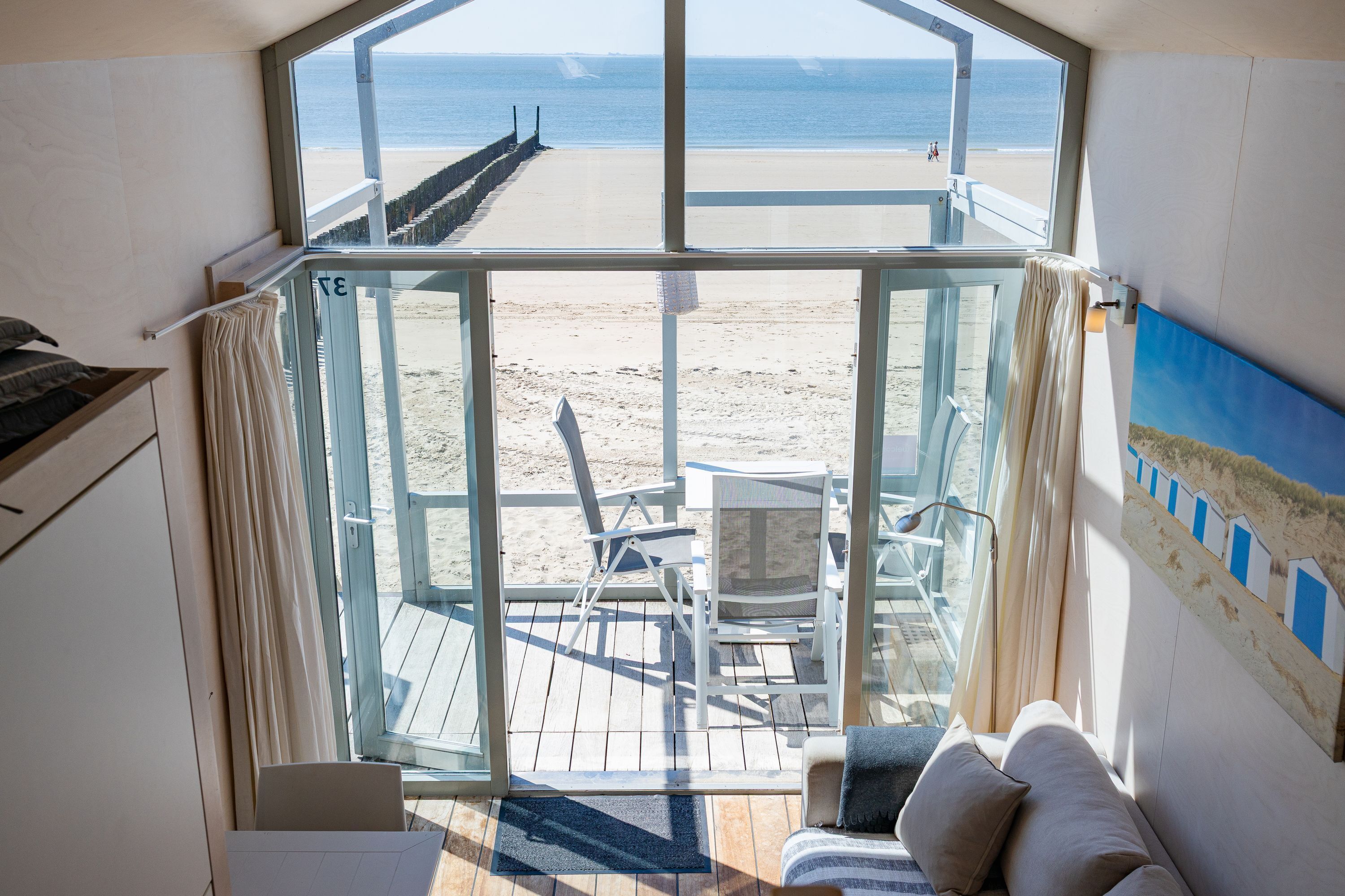 A room with a large glass door leading to a beach view, a white chair, a small table, and a sofa.