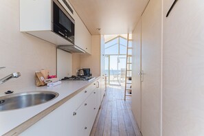 A narrow kitchen with a stainless steel sink, a stove, and cabinets. A wooden floor leads to a balcony with a view. - Slaapstrandhuisje - Strand Dishoek 37 I Dishoek (Vlissingen)