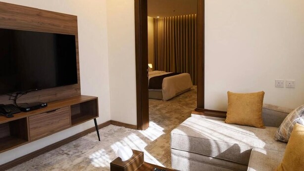 A hotel room with a TV on a wooden stand, a sofa with cushions, a small table, and a bed with curtains. - Hadaj Town Apartment Hotel (Al-'Ula)