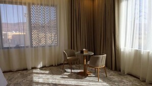 A room with a round table and two chairs, a patterned window blind, and a view of the outside. - Hadaj Town Apartment Hotel (Al-'Ula)
