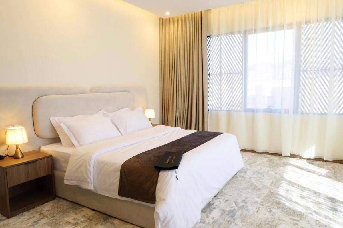 Una habitación de hotel con cama, mesita de noche, lámpara y ventana con cortinas.