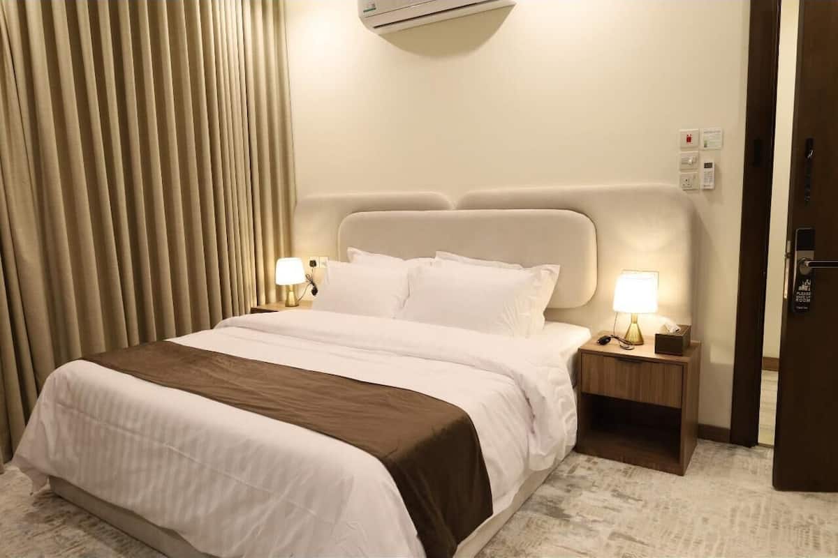 Una habitación de hotel con cama, lámparas de noche, mesita de noche, unidad de aire acondicionado, puerta y un aplique en la pared.