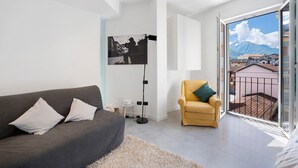 Ett modernt vardagsrum med en grå soffa, en gul fåtölj, ett inramat fotografi på väggen och en balkong med utsikt över byggnader och berg.