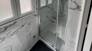 Eine gläserne Duschkabine mit Marmorwänden, eine schwarze Matte am Eingang und ein Fenster links.