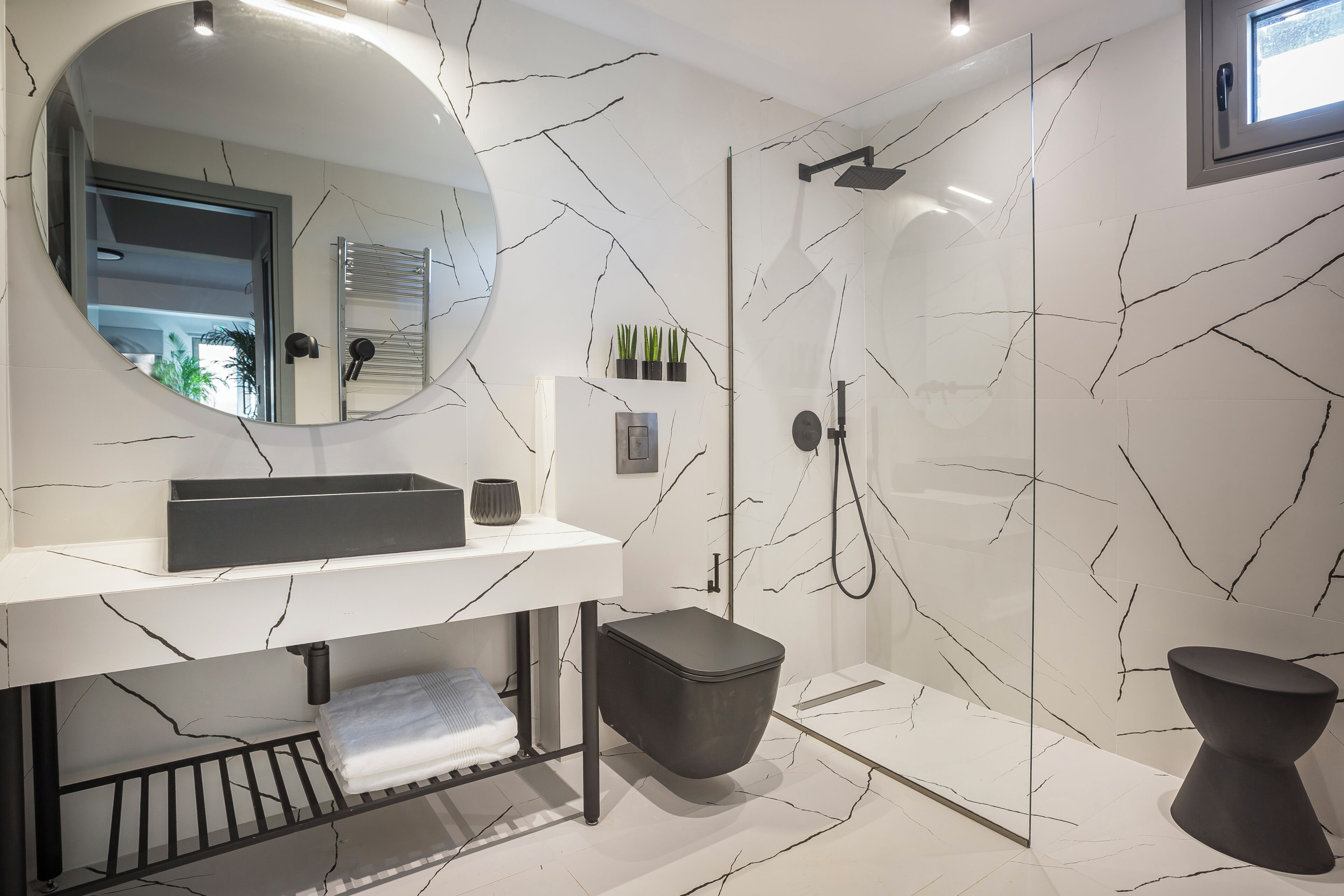 Un baño moderno con salpicadero de mármol blanco, un espejo redondo, un lavabo negro, un inodoro negro y una zona de ducha con mampara de cristal.
